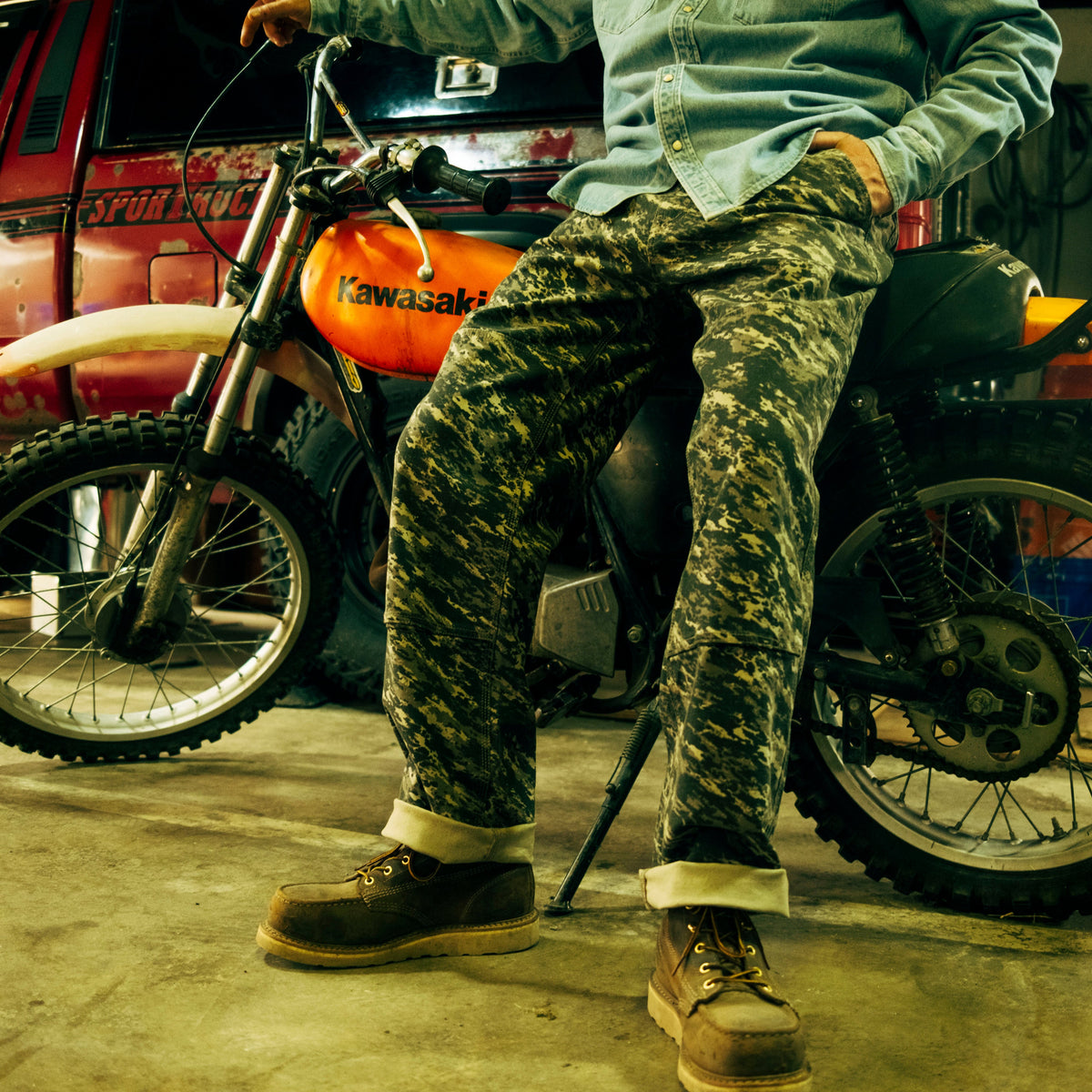General image of a man wearing camouflage utility pants and brown boots sitting on a Kawasaki motorcycle in front of a red truck.